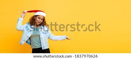 Fun waist up portrait of joyful African American woman wearing Christmas hat with open palm gesture on yellow isolated banner background with copy space