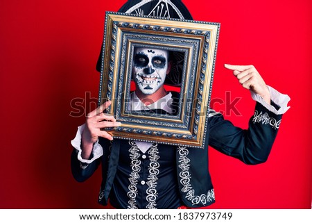 Young man wearing mexican day of the dead costume holding empty frame smiling happy pointing with hand and finger 