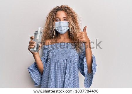 Beautiful caucasian teenager girl wearing medical mask holding hand sanitizer gel smiling happy and positive, thumb up doing excellent and approval sign 