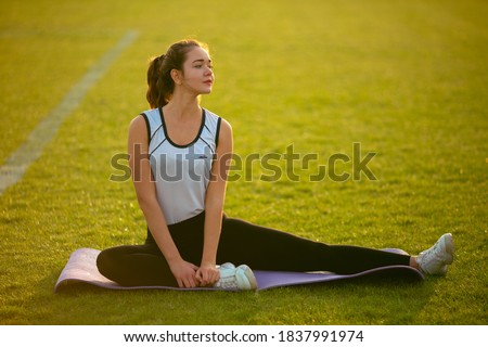 Fitness Girl Doing Leg Workout On Yoga Mat At Outdoor Stadium. Fit Woman With Strong Muscular Body In Fashion Sporty Outfit Doing Exercise Against stadium background. Active Lifestyle For Urban People
