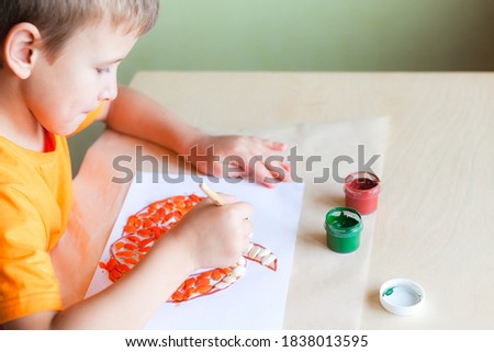 School boy making pumpkin from seeds on white paper, Halloween DIY concept. Step by step instruction. Step 3 Paint pumpkin seeds with orange color