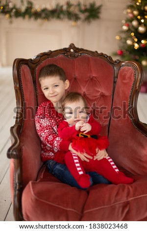 Litttle brother hugs his little sister in red retro chair near Christmas Tree. Enjoying a love hug, people's holidays. Togetherness concept.