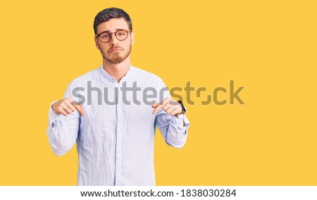 Handsome young man with bear wearing elegant business shirt and glasses pointing down looking sad and upset, indicating direction with fingers, unhappy and depressed. 