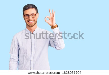 Handsome young man with bear wearing elegant business shirt and glasses smiling positive doing ok sign with hand and fingers. successful expression. 