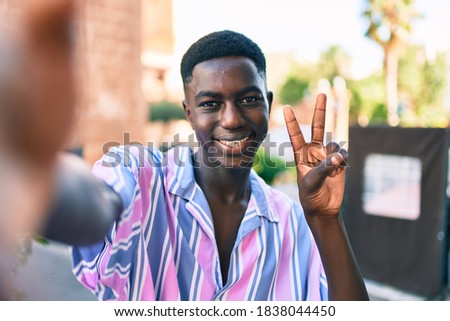 Young african american man making selfie by the camera doing victory sign with fingers at street of city.