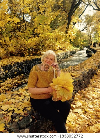 A beautiful blonde woman with a bouquet of yellow maple leaves sitting on stones on the bank of a mountain river against the background of trees with falling yellow foliage  Golden autumn landscape