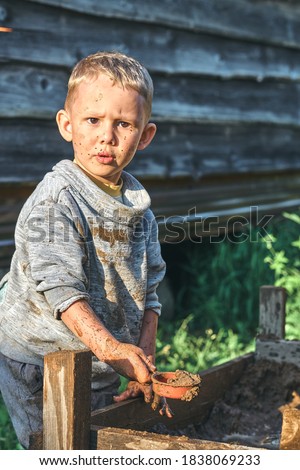 Cute serious preschooler boy with dirty face and clothes playing with toy cars in the sandbox near wooden rural house in summer day in countryside