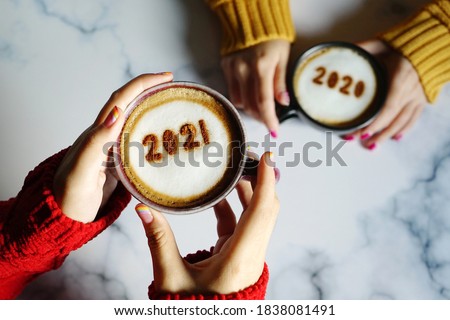 Goodbye 2020, Hello 2021 theme coffee cup with number 2021 on frothy surface in female hands holding and another one with number 2020 on frothy surface over marble table background. Holidays food art. Royalty-Free Stock Photo #1838081491