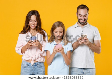 Smiling funny young parents mom dad with child kid daughter teen girl in basic t-shirts using mobile cell phones typing sms messages isolated on yellow background studio portrait. Family day concept