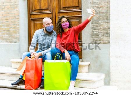 happy couple of a bearded bolt man and a curly caucasian woman wearing face mask carrying shopping bags sitting on stairs in the city center during black friday sales. girl and a boy taking a selfie