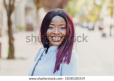 Portrait of a smiling young African American girl with pink hair and a blue shirt walking outside on a sunny day.