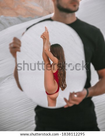 girl in red swimsuit is standing in gymnastic pose in reflection in round mirror in male hands
