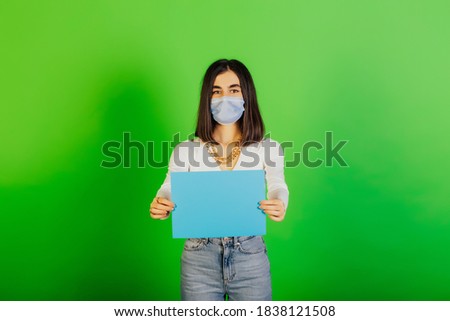 Serious girl wearing protective face mask holds empty blue paper sheet in hands, isolated on green background. Copy space.