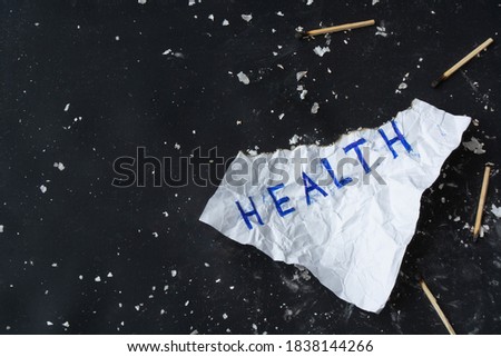 Crumpled burnt sheet of paper with inscription "Health" on black background with scattered ash and burnt matches . Health neglect and harm to health concept. Copy space. Top view