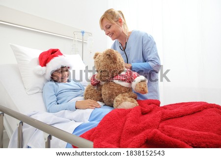 Christmas holiday in hospital happy child lying in bed with Santa Claus hat and nurse dressing a teddy bear 