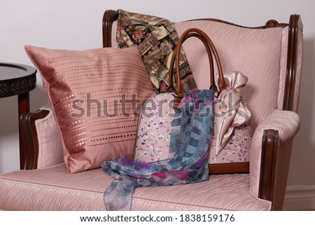In a family living room, the back of an antique pink chair, with two scarves, a satchel and a pink cushion, on a white wall background
