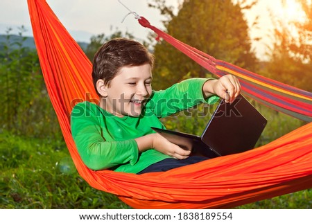 Boy in an orange hammock on a green background. The child is playing with a laptop. The concept of online learning in nature.
