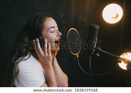 Young beautiful woman singing in microphone, recording voice in a studio with black walls and a lamp on the background.