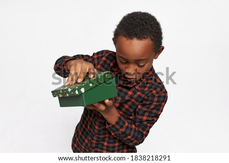 Isolated image of adorable African American boy expressing impatience, opening green cardboard box, curious to look inside and see present ahead of time. New Year, Christmas and winter holidays