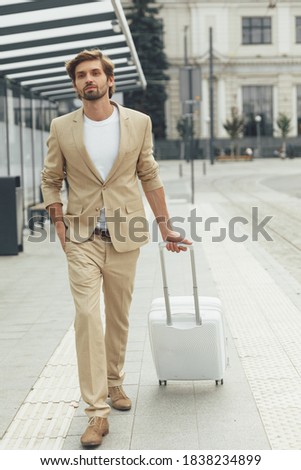Elegant young man with trendy hairstyle and beard dressed in business suit standing at bus stop with suitcase. Travelling concept. Business trip.