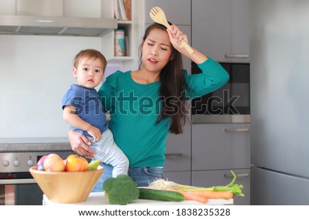 Stressed and tired mother with one hand carrying her baby while cooking at home kitchen.mixed race Asian-German family. Housewife over work, busy concept. Royalty-Free Stock Photo #1838235328