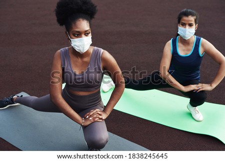 Two different ethnic girls stretch, stretch their muscles before training outdoors in a protective mask. Prevention of coronavirus