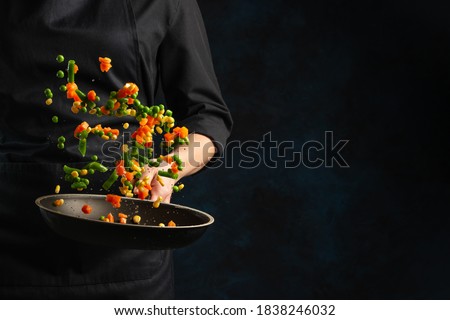 Professional chef in black uniform throws mix of frying corn, peas and asparagus in pan on dark blue background. Frozen motion. Backstage of cooking vegetarian dish. Creative preparing meal.