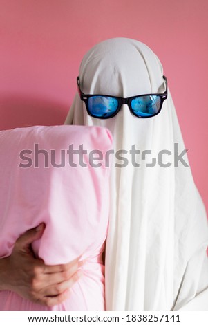 Cute sheet costume ghost with sunglasses hugging his pillow on a pink background. Halloween party carnival concept.