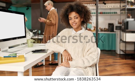 Creative female. A pretty young dark-skinned woman looking into a camera smiling while sitting on a chair at a table with her colleagues behind in a cosy creative workspace. Business concept