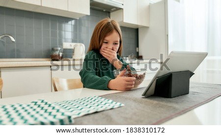 A littile girl is watching cartoons on her tablet and giggling while sitting alone at the table in a bright kitchen eating quick breakfast made of milk and cereal balls. Children and technology