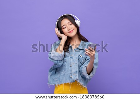 Happy Asian girl wearing headphones listening to music from mobile phone relaxing with eyes closed on isolated purple studio background