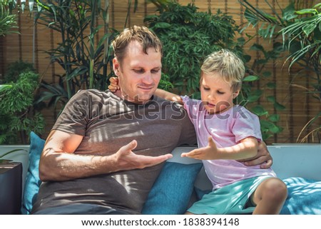 Father blond son sit look on open hands, play compare their palms. Child gently hugs daddy by neck. Bright sun beams light leaves garden terrace. Happy childhood, fatherhood love feelings, fathers day