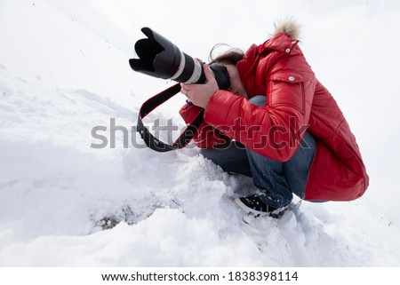 A photographer with a large lens takes pictures while sitting in the snow.