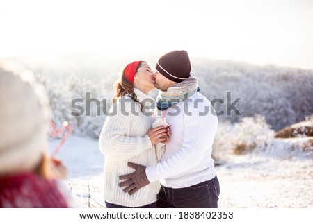 Preteen girl making shape of love heart standing  outdoors in snowy winter, at the background parents kissing. kid playing and have fun with the snow outside on season holidays