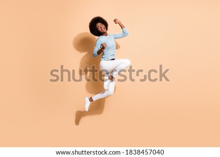 Photo portrait of glad african american woman jumping up holding fists up laughing isolated on pastel beige colored background