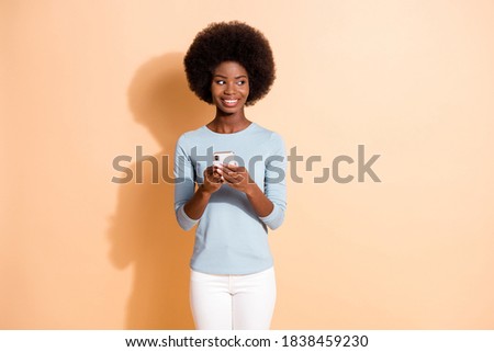 Photo portrait of happy smiling dark skinned girl keeping mobile phone searching looking at side isolated on beige color background