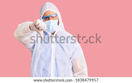 Young hispanic man wearing doctor protection coronavirus uniform and medical mask looking unhappy and angry showing rejection and negative with thumbs down gesture. bad expression. 