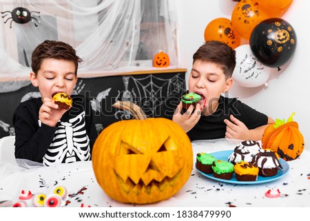 Kids having fun for halloween surrounded with scary decoration enjoying eating cupcakes. Jack O' Lantern Halloween pumpkin and cupcakes on the table. Happy Halloween!
