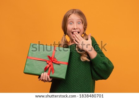 Image of charming blonde girl 12-14 years old in warm green sweater holding present box with red bow. Studio shot, yellow background, isolated. New Year Women's Day birthday holiday concept