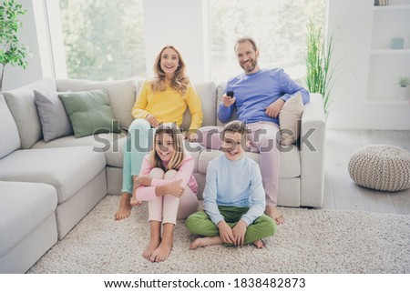Full length photo of comfort family watch tv hold remote control two small kids sit floor mom dad on couch in house indoors