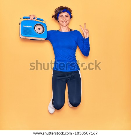 Young beautiful sportswoman listening to music smiling happy. Jumping with smile on face holding vintage radio doing victory sign over isolated yellow background.