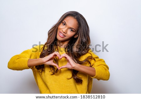 Lovely young african american woman with happy smile, shapes heart with both hands, expresses love to you, wears yellow wool sweater, says be my valentine flirts with boyfriend poses indoor copy space