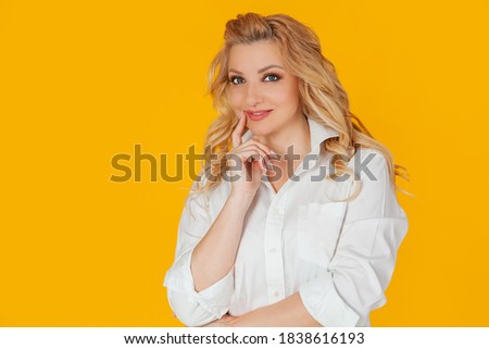Attractive happy European blond woman in white shirt smiling curiously, touching her finger to her lips thoughtfully. Standing on a yellow background