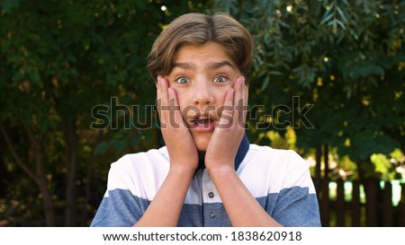 Close-up of the face of a young, cute, Caucasian boy who is surprised and puts the palms of his hands to his cheeks. Positive emotion. On the street. Royalty-Free Stock Photo #1838620918