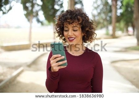Portrait of beautiful young woman with short curly hair wearing red T-Shirt, standing outdoors holding telephone being happy after receiving good and amazing news. 