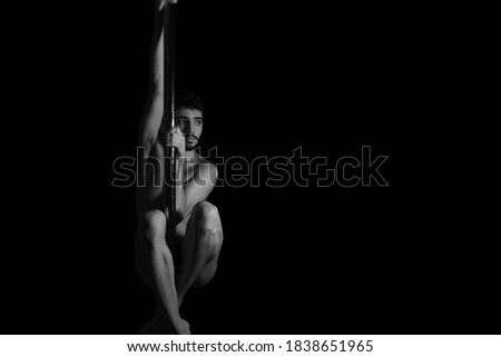 Dramatic dark portrait of a young man hanging from a pole dance bar with his eyes opened. Dark background studio shot. Black and white image.