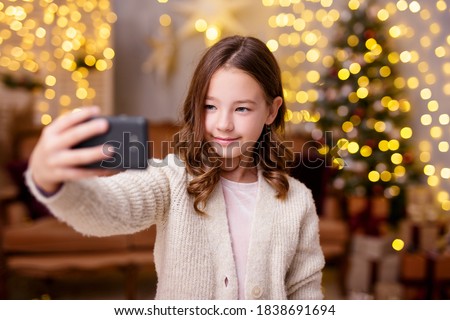Christmas and new year concept - cute little girl taking selfie photo with smartphone in decorated living room with Christmas tree and lights