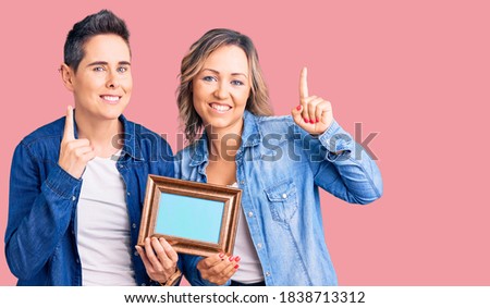 Couple of women holding empty frame pointing finger up with successful idea. exited and happy. number one. 