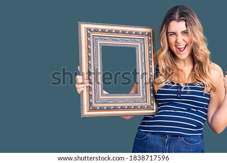 Young caucasian woman holding empty frame screaming proud, celebrating victory and success very excited with raised arms 