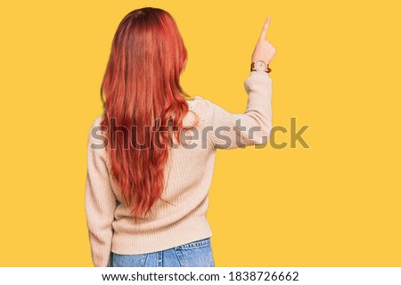 Young redhead woman wearing casual winter sweater posing backwards pointing ahead with finger hand 